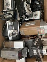 Top view of box showing multiple vintage cameras with leatherette and metal bodies, flash attachments, and camera cases