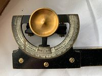 Close-up of the black metal inclinometer with semi-circular graduated dial, brass adjustment knob, and brass bell-shaped sight.