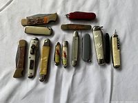 Group of twelve vintage miniature folding knives and tools laid out on white fabric. Varied in size, color and design.