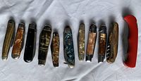 Photo shows 13 folding pocket knives, mostly vintage, with varied materials including bone, mother-of-pearl, plastic handles, plus one modern red plastic handled knife with serrated blade and corkscrew.