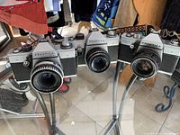 Three silver and black vintage Praktica 35mm film cameras on glass table in natural lighting showing front and lens sides.
