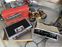 Lot of three vintage opera glasses on glass table: King brand with original red box, Novet pair with pearl shell case, and Lemaire Paris pair in brass finish.