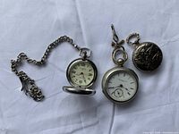Three vintage pocket watches arranged on a white cloth. One large silver-tone with chain and Arabic numerals, one medium silver-tone with Roman numerals and subdial, one small embossed silver-tone with a deer design on the case.