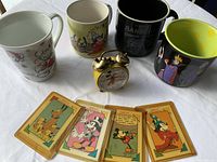 Photo of four Disney-themed mugs, four vintage Disney character playing cards, and a yellow Mickey Mouse alarm clock on white background.