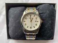 Men's Bulova 98B010 wristwatch with two-tone metal bracelet displaying time and date, shown on branded black pillow inside box.