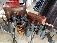 Two pairs of vintage binoculars with their brown leather cases placed on a glass table.