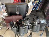Two pairs of black binoculars with carrying cases including one black and one brown leather case, plus an extra black case.
