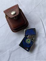 Photo showing a chrome Zippo lighter decorated with blue and yellow air force insignia and red US lettering on the front, alongside its original brown leather Zippo carrying case with snap button flap.