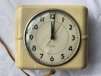 Front view of a vintage Westclox Belfast kitchen clock with cream square plastic casing and round clock face with black numerals and red second hand.
