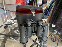 Front view of vintage Sersi binoculars showing textured black body, attached neck strap, and protective case behind with red interior lining.