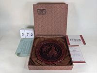 Plate displayed inside its original patterned box, resting on brown fabric with accompanying certificate and information sheet visible.