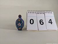 Oval-shaped cloisonné snuff bottle with floral patterns and metal stopper shown beside a number card.