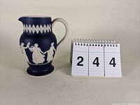 Front angled view of dark blue jasperware style Dudson Hanley pitcher decorated with white classical dancing figures and floral bands with measuring cards nearby.