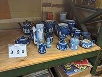 Photo showing group of over 15 blue jasperware pieces in Wedgwood style with white classical relief decoration arranged on a wooden shelf.