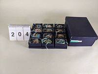 12 cloisonné egg ornaments displayed in individual compartments in a blue box with a small calendar showing number 204 beside them.