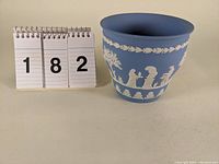 Front and side views of light blue Wedgwood jasperware cache pot showing white classical relief decoration and flared rim.