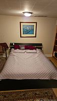 Front view of wood and metal bed frame with gray quilted blanket, multiple cushions, framed painting on wall and bookshelf in background