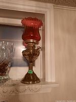 Full view of brass oil lamp with red glass shade on a window sill, showing base and shade.