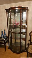 Front and side view of the wood framed glass curio cabinet with glass shelves and doors. Shows interior lighting and curved glass panels. Cabinet is empty except for some decorative items not included.