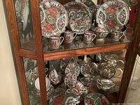 Overview of the china set displayed in a glass cabinet, showing plates, cups, and bowls with intricate floral and figure patterns.