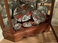 Wide angle photo showing the entire 7-piece porcelain set displayed inside a wooden cabinet, showcasing platter, plates, cups, covered bowl, and sugar bowl with the three wooden stands.