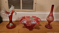 Three cranberry glass pieces displayed side by side on a wooden floor with heating vent in background. Items include horn-shaped vase, ruffled bowl, and tall slender vase.