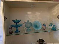 Full shelf view showing all glass items including blue candy dishes, purple floral vase, clear decanter, blue bowl, egg shaped piece, and white basket.
