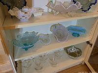 Four decorative glass bowls displayed inside a white shelf cabinet including blue floral embossed bowls and white ruffled-edge bowls.