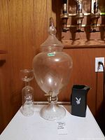 Photo showing large etched glass jug with brass spout, small glass jug with attached cup, and black plastic Playboy pitcher on white surface against wooden wall.