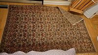 Top-down view of full 6'x9' rectangular oriental area rug with floral and geometric design in brown, beige, red, and black tones. Corner folded to show backing.