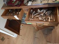Top view of wooden cabinet drawers containing assorted silver plate cutlery and serving utensils.