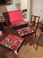 Three black metal cases with red felt lining containing assorted flatware on wooden table, with silver pedestal stand.
