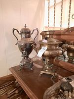 Two urns: silver-plated and brass, displayed on a wooden surface with cutlery beneath them.