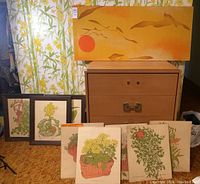 Photo showing four framed prints and five unframed botanical art pieces arranged in front of a chest, with large stretched canvas leaning above.