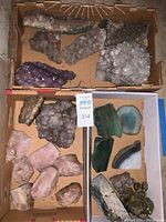 Overview of multiple mineral and stone specimens in cardboard boxes including amethyst clusters, quartz, rose quartz, agate slices and pyrite pieces.
