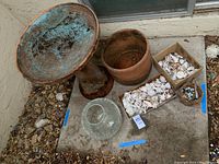 Overview of large terra-cotta birdbath with blue enamel interior, terra-cotta planter, clear crackle glass vase, and containers of assorted seashells on outdoor concrete floor.