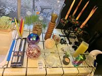 Overview of eclectic candle and candlestick collection on tiled surface, showing tapered candles, glass spheres, yellow block candle, wooden candlestick, and other decorative items.