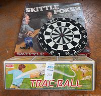 Overview photo showing the Skittle Poker game box, the round Marksman dart board on top, and the Trac-Ball racquet game box in front.