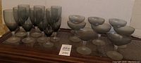 Photo of 9 taller, tinted Fostoria water goblets on left and 8 champagne coupes with frosted bowls on right, arranged on wood surface.