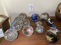Photo showing multiple clear glass ball paperweights with air bubbles, blue art glass paperweight, black and gold leaf paperweight, star-shaped paperweight, and iridescent heart-shaped paperweights.