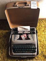 Front angled view of Smith Corona manual typewriter inside beige carrying case showing keyboard with green keys, typed sheet, and ink ribbon box.