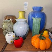 Photo showing six handmade blown glass vases and three decorative glass fruits on wooden surface against beige wall.