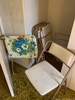 Four stacked vintage metal folding chairs with white vinyl seats and backs plus one floral designed metal TV tray with folding legs.