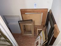 Various wood frames and stacked unused white canvases leaning against a white wall on wooden floor, some small matted artworks beside them.