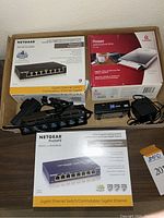 View of the boxed NETGEAR 8-port Gigabit Ethernet Switch, boxed IOmega USB powered floppy drive, and smaller USB devices including StarTech.com hub and Linksys Cisco switch beside power adapters.