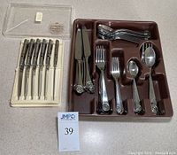 Plastic tray holding Robert Welch Ammonite flatware service for 8 with knives, forks, and spoons. Next to the tray is a clear box holding six Briddell Carvel Hall steak cutters with cream handles showing deterioration.
