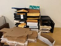 Stack of envelopes and folded cardboard folder organizers in front of black vertical letter trays and binders on a table