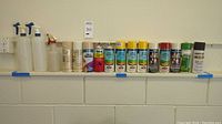 Photo showing the entire line of assorted spray paint cans and spray bottles on a shelf against a wall, to document lot contents and amount.