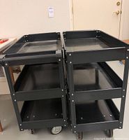 Two black steel rolling carts side by side, showing three levels each and caster wheels.