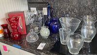 Wide view of all glass vases and decor arranged on countertop including red Lenox vase boxed, clear vases, cobalt blue decanter and vase, and iridescent glass marbles